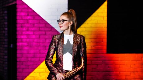 A woman stands in front of a coloured wall with an alto saxophone