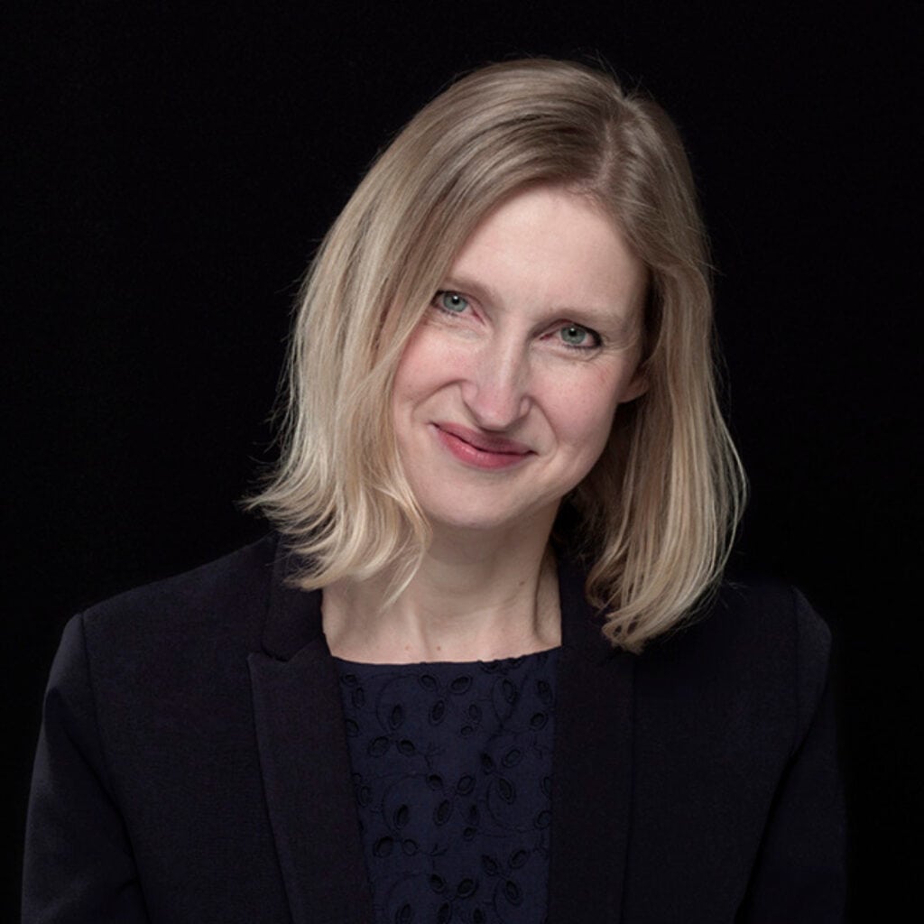 Historian Tracy Borman stands against a black background looking at the camera.