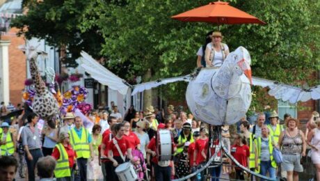 A giant model bird, ridden by two adults, leads a parade down the street