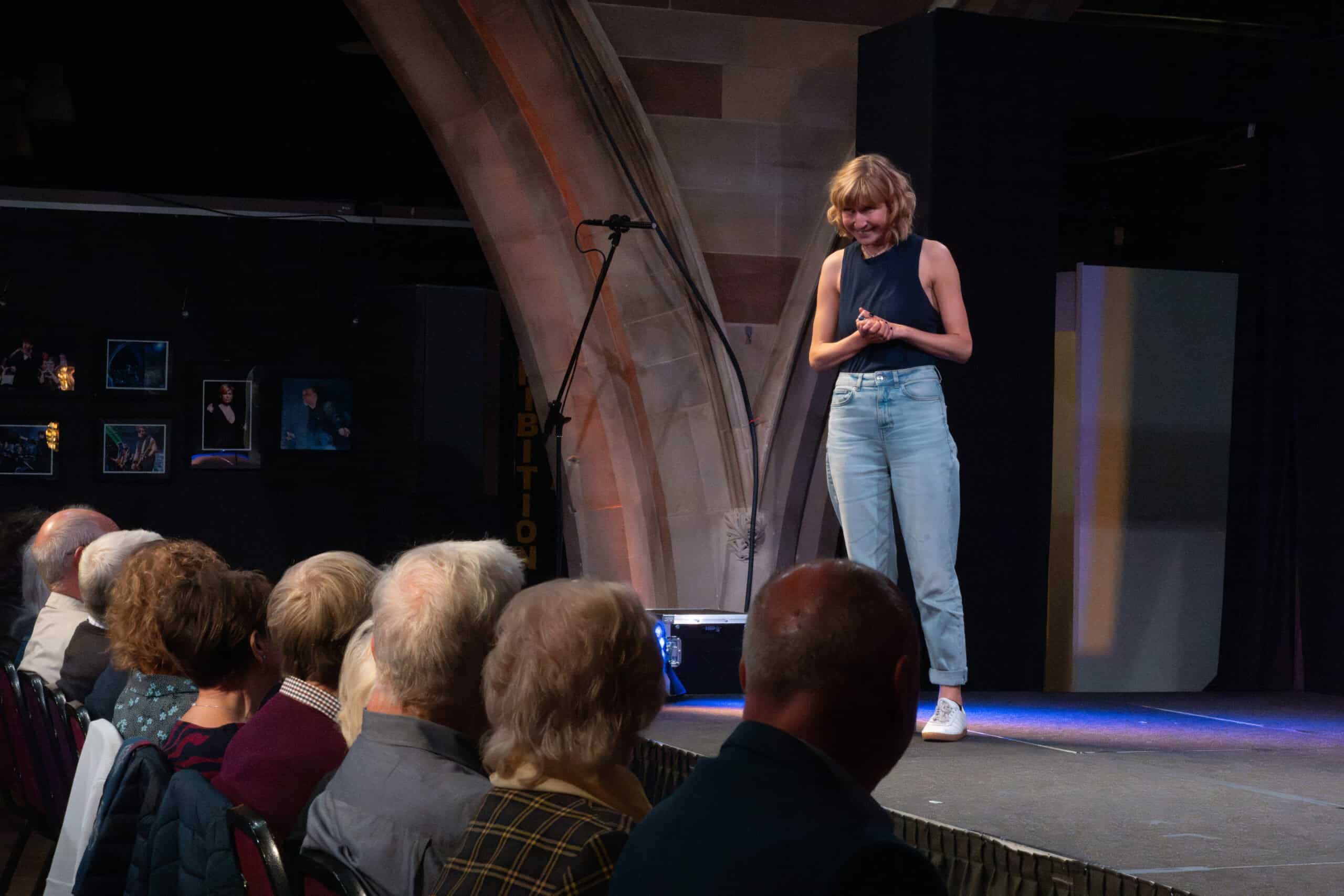A performer stands on stage talking to audiences with their hands raised.