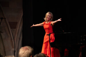 Liza Pulman stands on stage, arms open wide and smiling, wearing a red dress