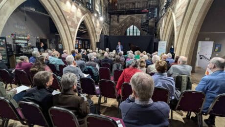 A view from behind of audience members sat in rows in a converted church, as Festival Director Damian Thantrey speaks.