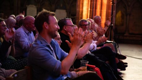 A lively audience applauds during a concert at Lichfield Festival in a historic, stone-arched setting.