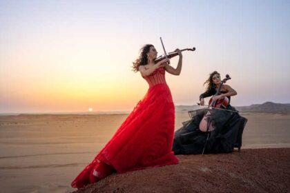 Two women stand in a desert playing a violin and cello