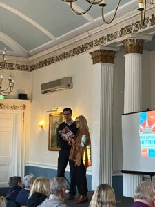 Damien Thantrey and Anne Gaithwaite stand on a stage in a white room reading from a book