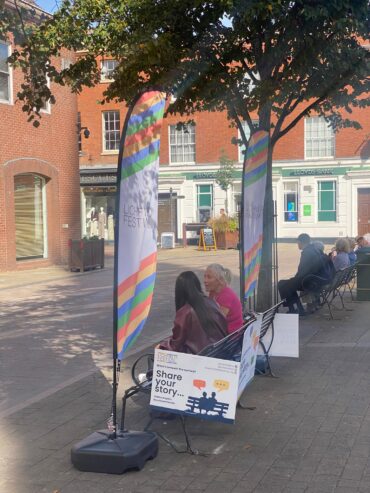 A man and woman sit on a bench with a microphone surrounded by Festival branding