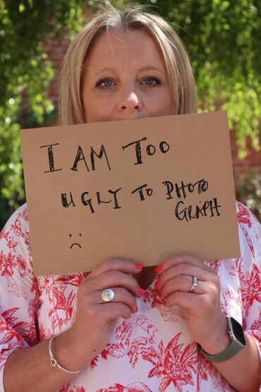 A woman holds a cardboard sign that says 'i'm too ugly to photograph'